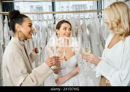 Two brides in wedding attire and a woman with champagne flutes in front of a rack of wedding dresses in bridal salon. Stock Photo