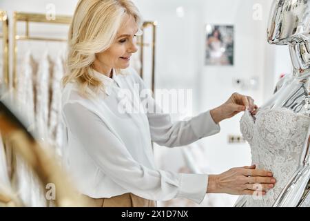 Middle-aged beauty shop assistant admires wedding dress on hanger in bridal boutique. Stock Photo