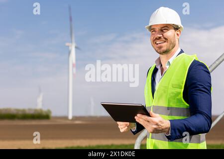 A cheerful engineer in safety gear reviews data on a tablet before towering windmills under a clear sky. Stock Photo