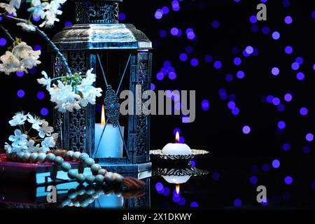 Arabic lantern, Quran, misbaha, burning candle and flowers on mirror surface against blurred lights at night. Space for text Stock Photo