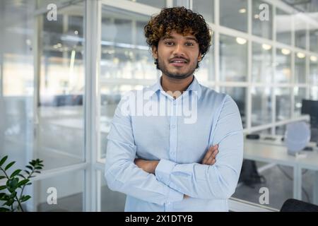 A portrait of a confident young businessman with curly hair, standing arms crossed in a light-filled, contemporary office environment. Stock Photo