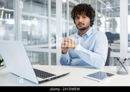 Confident male customer support representative with curly hair, using a headset, works on a laptop in a modern office setting. Stock Photo
