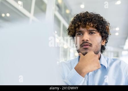 A young curly-haired man dressed in a blue shirt, pondering deeply while sitting in a bright, contemporary office environment. His expression reflects concentration and contemplation. Stock Photo