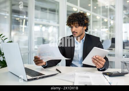 Focused young professional businessman reviewing financial documents while using a laptop and tablet in a modern, bright office space. Stock Photo