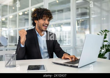 A joyful young man sits in a modern office, celebrating a successful moment with his hand raised in victory while using a laptop. Stock Photo