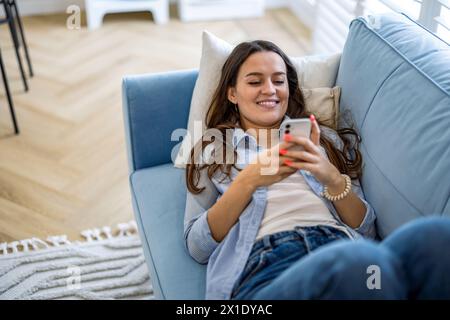 Smiling woman using mobile phone on sofa in living room at home Stock Photo