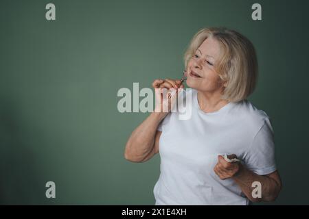 Senior woman doing skin care treatment using face roller isolated over green background with copy space. Beauty treatment, skin care, wellbeing. Stock Photo