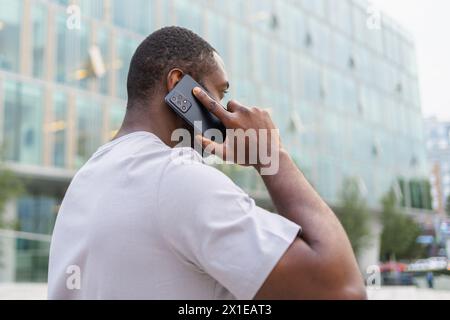 Back view African American man talking on smartphone outdoor. Unrecognizable person guy with cell phone walking on street in city. Man making answering call by cellphone having conversation by mobile Stock Photo