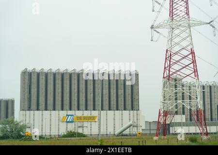 Ghent, Belgium - May 22, 2023: A tall lattice high voltage tower stands beside a large industrial facility with multiple silos, under a cloudy sky. Stock Photo