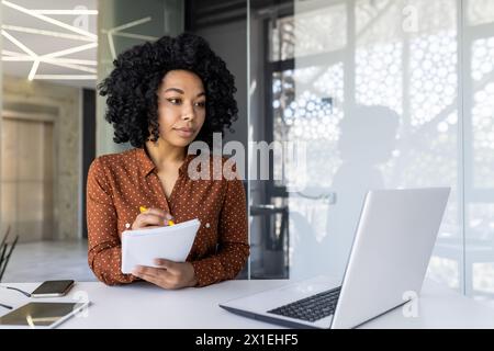 A focused young African American woman working diligently in a bright, modern office space, taking notes while consulting her laptop. Stock Photo