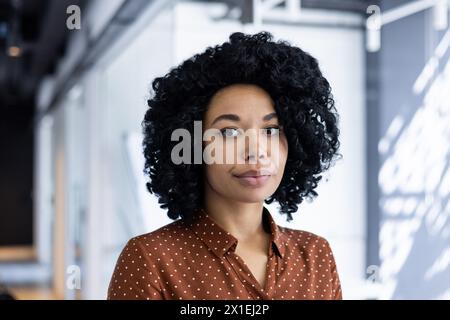 A young African American businesswoman with curly hair, wearing a polka dot blouse, stands confidently in a well-lit modern office environment, exuding professionalism and poise. Stock Photo