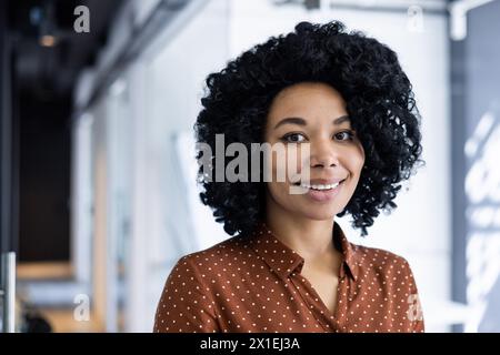 Portrait of a confident young African American businesswoman smiling warmly, dressed in a stylish polka dot blouse in a modern office setting. Stock Photo