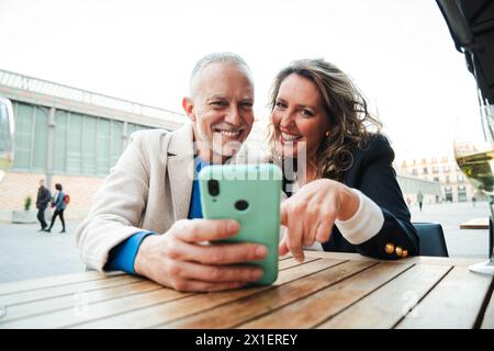 Middle aged couple having fun sharing media with a cellphone. Mature people watching the smartphone on a coffee shop, restaurant or bar. Mid adult friends buying on a market place using a mobile phone. High quality photo Stock Photo