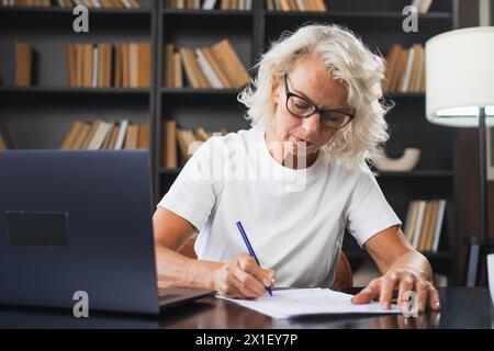 Middle aged senior woman using laptop computer writing notes at workplace. Focused mature old businesswoman work at office making written records doing online work. Mature boss professional worker Stock Photo