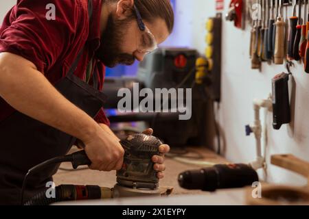 Carpenter wears protective glasses at workbench, using orbital sander with fine sandpaper on lumber to achieve refined finish. Man in assembly shop uses angle grinder on wood for professional results Stock Photo