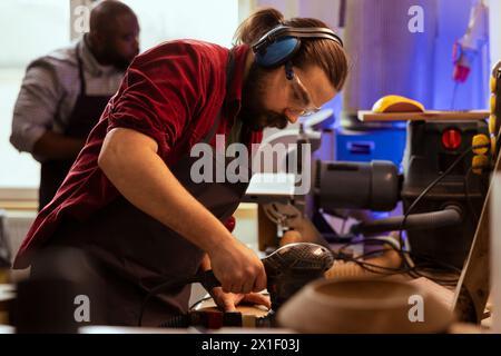 Artisan in joinery wearing safety earmuffs using orbital sander on lumber. Man in carpentry shop uses angle grinder to create wooden objects helped by colleague in blurry background Stock Photo