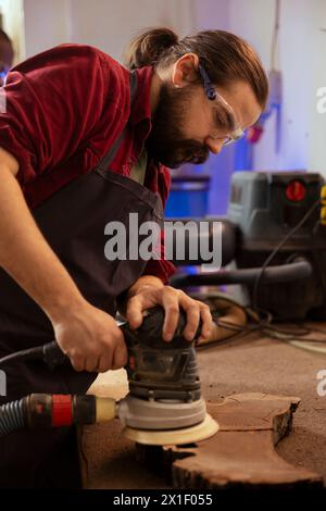 Cabinetmaker wearing protection glasses using angle grinder on wood to smooth surfaces by abrasion with sandpaper. Expert using orbital sander gear for furniture assembling job in carpentry shop Stock Photo