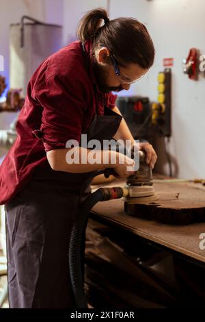 Craftsperson at work using orbital sander with fine sandpaper on lumber to achieve smooth finish. Woodworking expert in carpentry shop uses angle grinder on wood for professional results Stock Photo