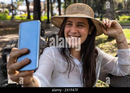 In spring young Latin American woman (33) is sitting on a park bench smiling while using her cell phone for a self-portrait. while using her cell phon Stock Photo