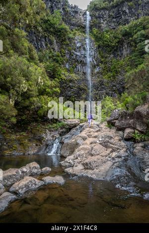 Description: Female tourist with backpack walks over rocks at waterfall falling from high rock wall. Lagoa do Vento waterfall, Madeira Island, Portuga Stock Photo