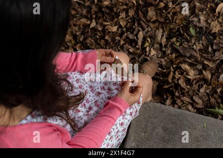 Top view of young Latin American woman (33) holding a dry leaf in her hands. She is wearing a flower dress and a pink sweater. Background of dry leave Stock Photo