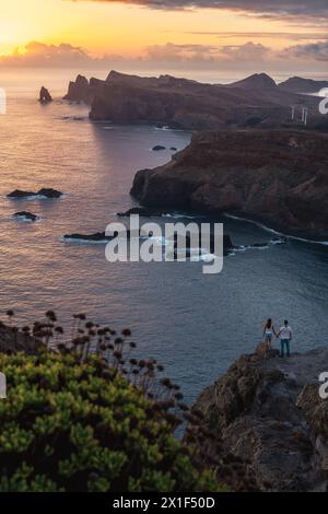 Description: Tourist pair enjoys panoramic sunrise from a vantage point on a steep cliff over the seascape and along the rugged foothills of the Madei Stock Photo