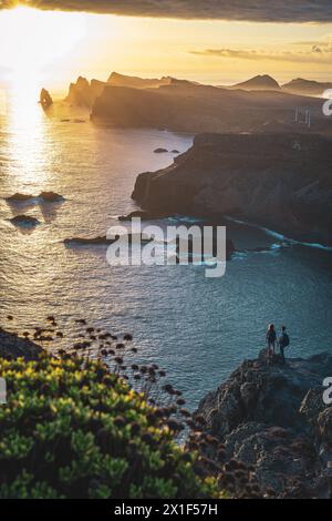 Description: Tourist couple with backpacks enjoys sunrise from vantage point on steep cliff above seascape and along rugged foothills of Madeira's coa Stock Photo