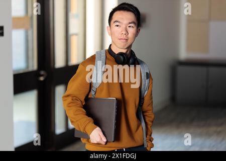 A young Asian college student is walking down a hallway in school holding his laptop Stock Photo