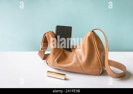 Woman leather handbag with phone and lipstick. On white table and blue background, copy space. Stock Photo
