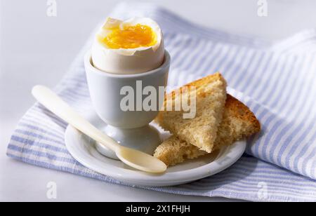 Boiled egg with top cut off in an eggcup, two slices of toast and a spoon on a striped napkin. Stock Photo