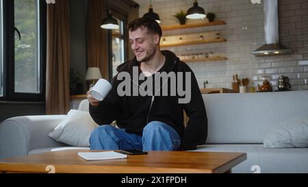 Funny guy sitting on the couch looking at the camera, talking, showing the mug, drinking coffee or tea, enjoying his weekend free time at home. Stock Photo