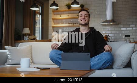 Peaceful smiling young man sitting in lotus pose on the couch and meditating taking deep breaths. Stock Photo