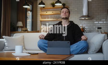 Peaceful young man sitting in lotus pose on the couch and meditating taking deep breaths. Stock Photo