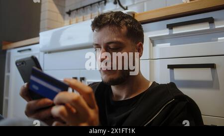 Young man sitting on the kitchen floor with a credit card and smartphone in hand enters credit card number for online shopping, making electronic paym Stock Photo