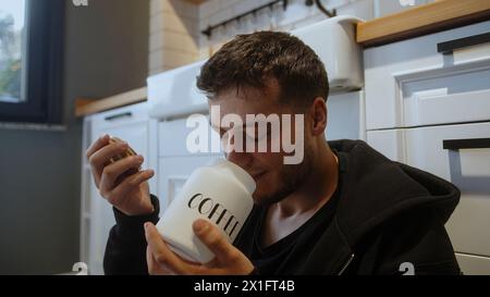 Young man sitting on the kitchen floor holds a jar of coffee, opens, smells the coffee and smiling. Stock Photo