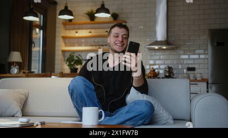 Smiling man sitting on sofa holding mobile phone, waving and greeting while having video conference call on smartphone. Man looking at screen, talking Stock Photo