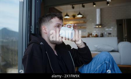 Young man sitting on the floor leaning against the balcony door, drinking morning coffee or tea from a mug. Enjoying leisure time at home Stock Photo