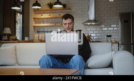Man working on laptop computer while sitting on a sofa in living room. Browsing internet, drinking coffee from a mug, having fun at home. Stock Photo
