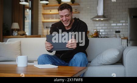 Overjoyed man playing a video game on a tablet computer while sitting on the sofa at home. Excited concentrated man engrossed in interesting game, hav Stock Photo