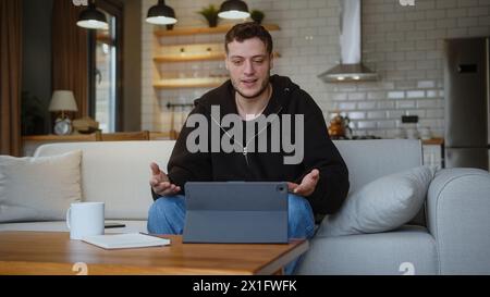 Smiling man sit on the sofa having video call conversation through using tablet computer at home. Share news, talk to family or friends, enjoy online Stock Photo