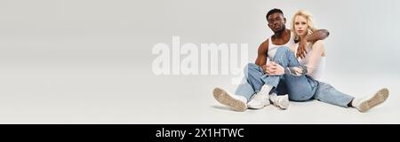 A man and a woman, a young multicultural couple, peacefully sit on the ground in a studio against a grey background. Stock Photo