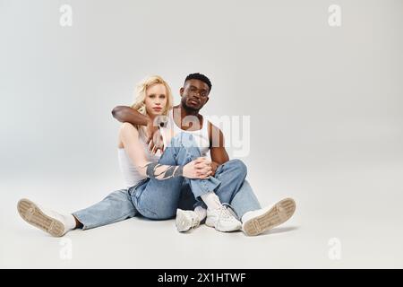 A young man and woman sit closely on the ground, lost in conversation and connection against a grey backdrop. Stock Photo