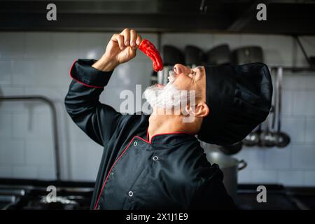Mature chef in uniform playfully tastes a fresh red chili pepper in a commercial kitchen Stock Photo