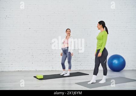 Two women, one pregnant, stand on yoga mats in a gym, engaged in a serene moment of mindfulness and movement during a parents course. Stock Photo
