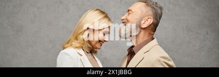 An elegant, mature loving couple in debonair attires pose together on a gray backdrop. Stock Photo