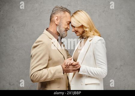 Mature man and woman, elegant in debonair attire, stand side by side in a loving pose on a gray backdrop. Stock Photo