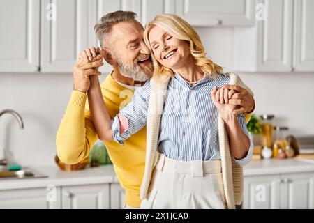 A mature loving couple dressed in cozy homewear dancing joyfully in their kitchen at home. Stock Photo