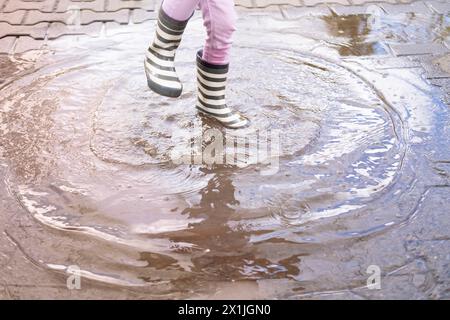 closeup 5-year-old girl joyfully jumps in puddle wearing rubber boots, children's feet in splashes water, happily jumping, pleasures of childhood Stock Photo