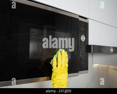 A girl's hand in a yellow glove with a sponge washes the microwave oven from grease and dirt. Foam and anti-grease. Stock Photo