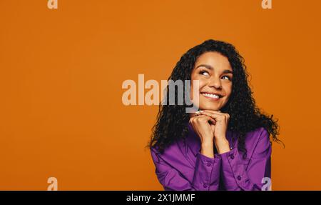 On a orange background, a cheerful woman with curly hair and a vibrant smile gazes thoughtfully. Her colorful attire adds to her youthful and casual v Stock Photo
